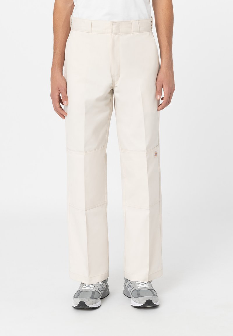Pantalones de tiro ancho en color beige con una textura suave, que cuentan con paneles cosidos y una etiqueta de marca visible en el lateral, combinados con zapatillas gris.