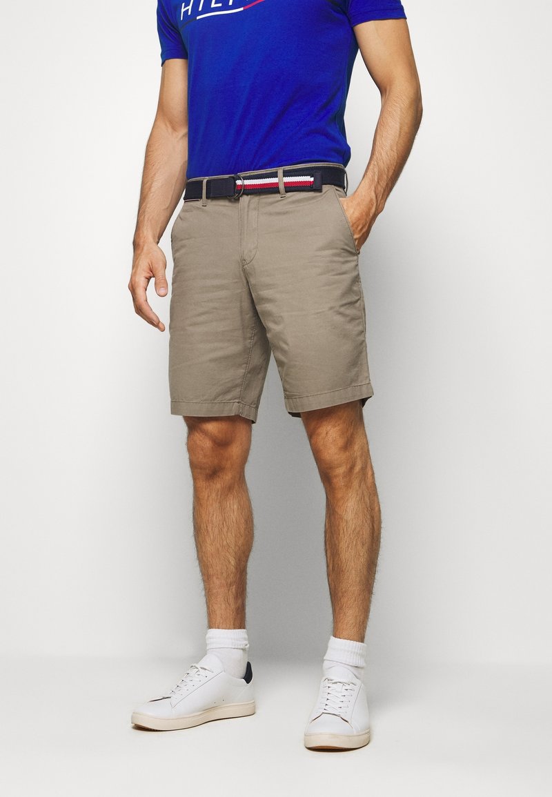 Hombre que lleva pantalones cortos beige con un cinturón a rayas, camisa azul, calcetines blancos y zapatillas blancas, de pie con una mano en el bolsillo.