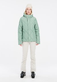 Mintgrüner, gepolsterter Mantel mit Rautensteppung, Kapuze und Druckknöpfen vorne; getragen mit cremfarbenen Hosen und schwarzen Skischuhen.