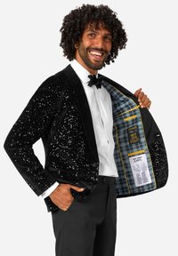 Moški se smeška v črnem bleščečem sakou, beli srajci in metuljčku, ki prikazuje modro karo podlogo z oznakami znotraj jakne.
