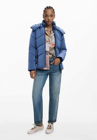 Blå dunjakke med hætte og quiltet design, lagdelt over en mønstret skjorte, kombineret med lyseblå jeans og beige sko.