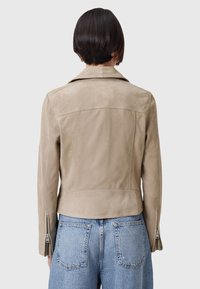Beige suède jack met een getailleerd ontwerp, schouderkraag en ritssluiting aan de mouwen, weergegeven vanuit de achterkant tegen een effen achtergrond.
