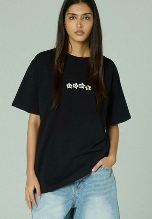 Joven con cabello largo y oscuro, vistiendo una camiseta negra holgada con el texto "GRMY" y jeans de mezclilla azul claro, parada frente a un fondo neutral.