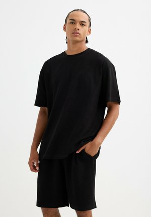 Joven con el cabello trenzado, de pie con una mano en el bolsillo, vistiendo una camiseta negra holgada y pantalones cortos negros sobre fondo blanco.