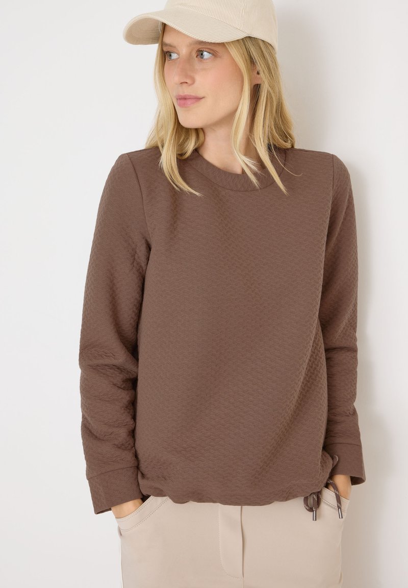 Felpa marrone con texture, maniche lunghe e scollatura rotonda, con orlo arricciato, abbinata a un cappellino chiaro.