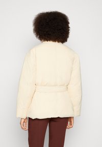 Casaco puffer em tom creme com cintura cingida, ombros arredondados e um acabamento texturizado. Apresenta uma silhueta justa e mangas longas.