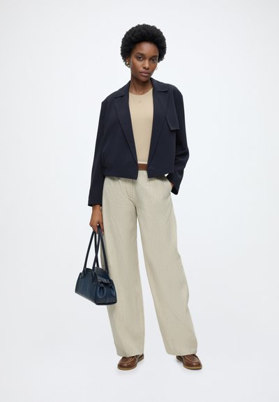 Femme aux cheveux courts et bouclés portant un blazer noir, un haut beige, un pantalon large clair, des chaussures marron, tenant un sac à main bleu foncé.