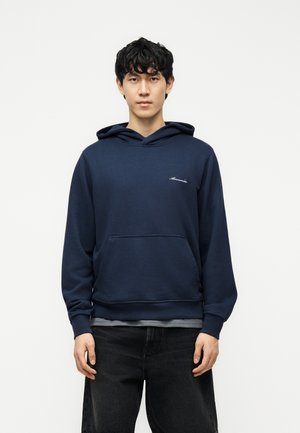 Junger Mann mit schwarzen lockigen Haaren, der einen marineblauen Kapuzenpullover mit Fronttasche und dunkle Hose trägt, vor einem schlichten weißen Hintergrund steht.