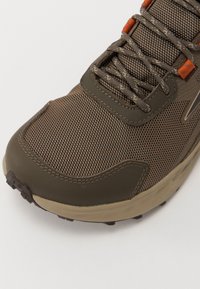 Brun idrettssko med mesh-overdel, gummisåle, broderte detaljer og oransje snøresløyfer. Teksturert finish og avrundet tådesign.