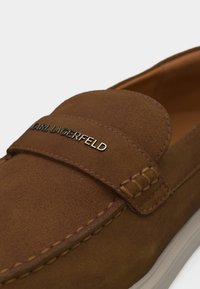 Καφέ μοκασίνι από καστόρι με λευκή σόλα και μεταλλικό λογότυπο "KARL LAGERFELD" στο λουράκι μπροστά.