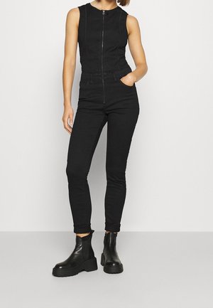 Woman wearing sleeveless black zip-up jumpsuit with slim fit pants and black chunky ankle boots, standing with one hand in pocket.