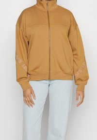 Sweatshirt zippé marron avec un col haut, présentant une texture lisse et un logo en relief sur les manches. Associé à un pantalon en jean clair.
