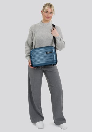 Blauwe hard-shell schoudertas met ribbelontwerp, zwarte accenten, verstelbare schouderband, vastgehouden door een persoon in grijze kleding, tegen een lichte achtergrond.