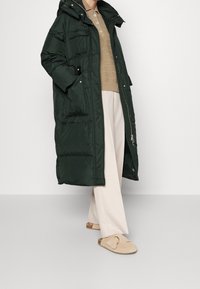 Lang, mørkegrøn pufferjakke med hætte, der har flere lommer og lynlås lukning, båret over en beige strikket sweater og lyse bukser.