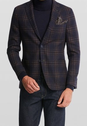 Uomo che indossa un blazer a quadri blu navy scuro e marrone sopra un dolcevita blu navy, abbinato a jeans blu scuro e un fazzoletto da taschino a fantasia.