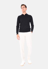 Uomo in camicia nera a maniche lunghe, pantaloni bianchi e scarpe bianche, in piedi con una mano in tasca contro uno sfondo chiaro e semplice.