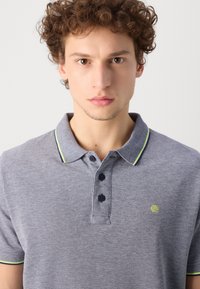 Camisa polo de color azul marino con un tejido texturizado, tres botones y acentos en verde contrastante en el cuello y las mangas. Pequeño logo en el pecho.