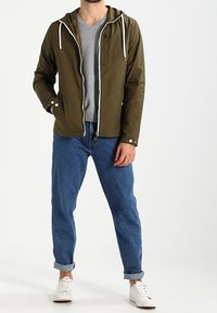 Svetle olivově zelená lehká bunda s kapucí a bílými stahovacími šňůrkami, předními kapsami, v kombinaci se světle šedým tričkem s výstřihem do V a modrými ohrnutými džíny.