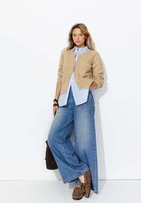 Gilet en maille beige porté sur une chemise rayée bleue et blanche, associé à un jean en denim à jambes larges et des chaussures à imprimé léopard. Un sac marron complète le look.