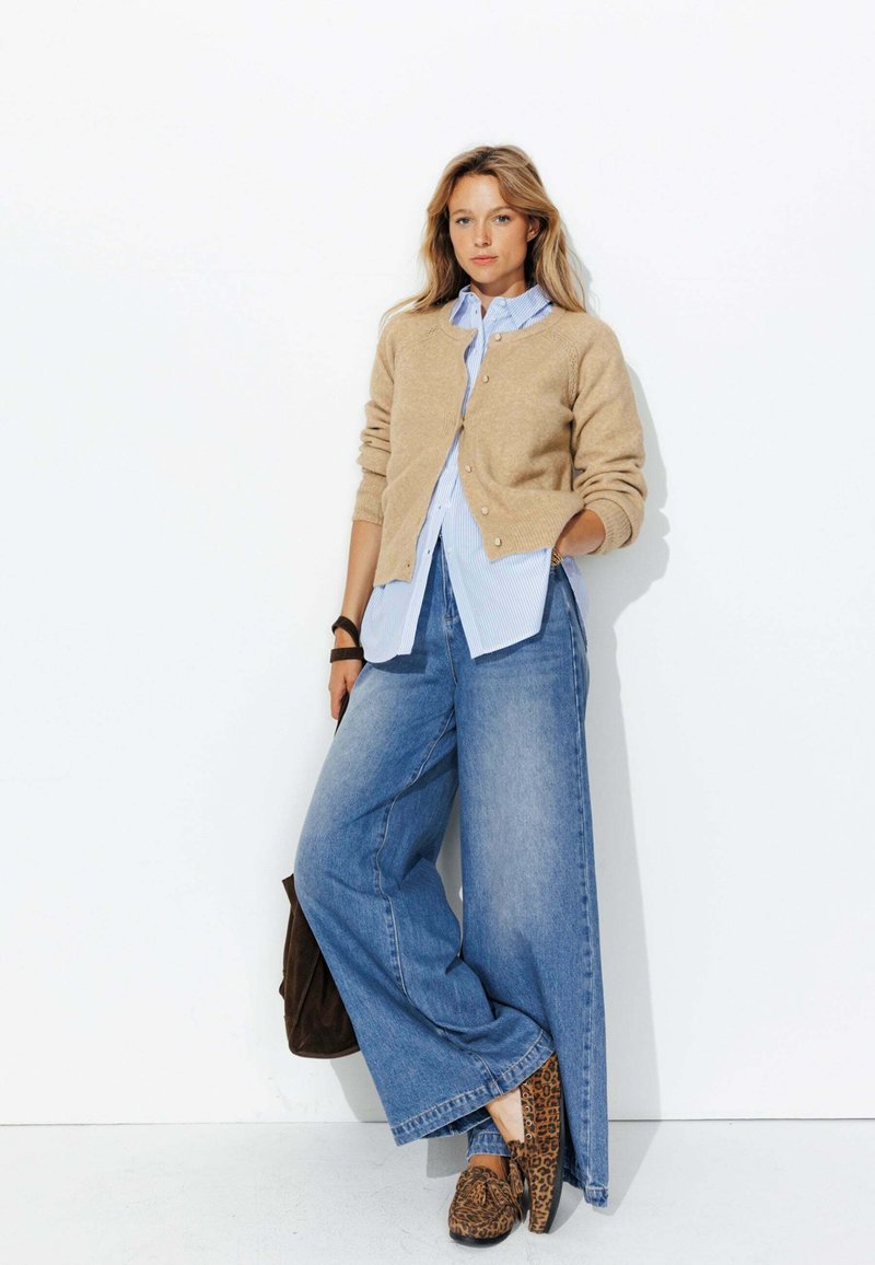 Gilet en maille beige porté sur une chemise rayée bleue et blanche, associé à un jean en denim à jambes larges et des chaussures à imprimé léopard. Un sac marron complète le look.