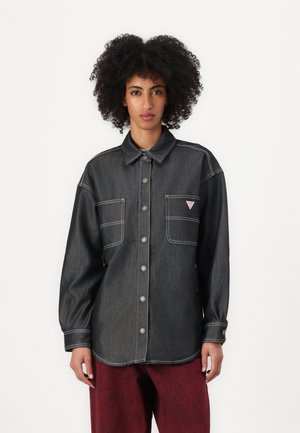 Mørk denimskjorte med knapfront, to brystlommer, kontrasterende hvit søm og et lite logo-patch.