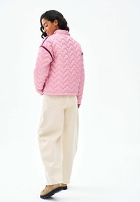 Žena otočená chrbtom, oblečená v prešívanej ružovej bunde, širokých béžových nohaviciach a svetlohnedých topánkach na bielom pozadí.