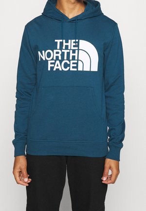 Mężczyzna ubrany w turkusową bluzę z kapturem z białym logo The North Face z przodu i czarne spodnie, stojący na jednolitym białym tle.