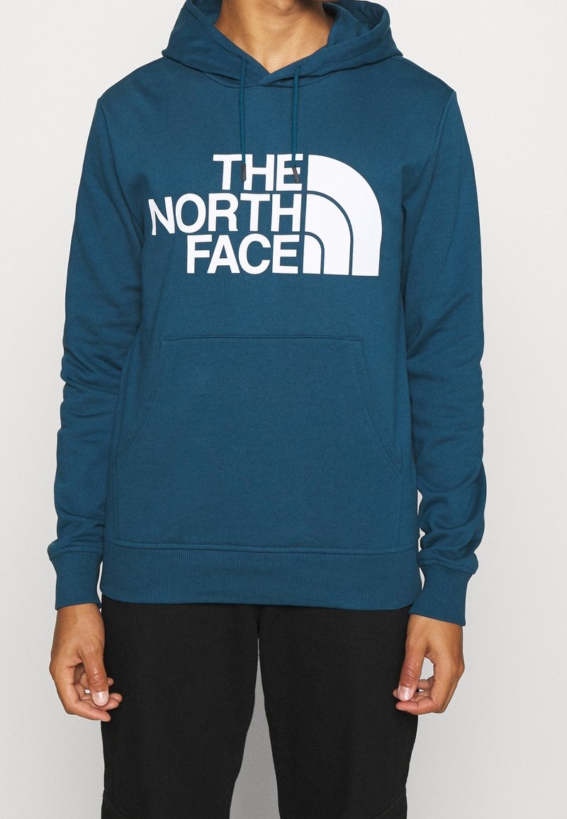 Homme portant un sweat à capuche bleu sarcelle avec un logo blanc The North Face sur le devant et un pantalon noir, debout devant un fond blanc uni.