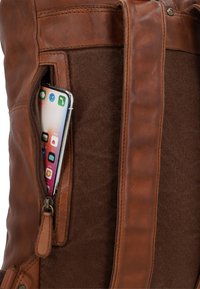 Brauner Leder- und Stoffrucksack mit einer teilweise geöffneten Seitentasche, die ein Smartphone mit App-Symbolen auf dem Bildschirm hält.