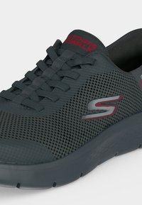 Тъмно сив маратонка Skechers без връзки с мрежест горен материал, детайли с връзки и червено и сиво лого "S" отстрани.