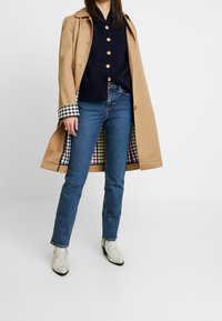 Gabardina beige con forro de cuadros en blanco y negro, suéter de punto azul marino, jeans azules y botas tobilleras blancas con estampado de serpiente.