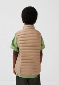 Beige donsbodywarmer met horizontale stiknaden,staande kraag en mouwloos, gedragen over een groen t-shirt. Eenvoudig, functioneel ontwerp.