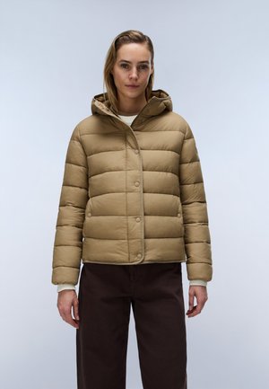 Vrouw draagt een beige gewatteerde jas met capuchon en donkere broek, staand tegen een effen lichtgrijze achtergrond.