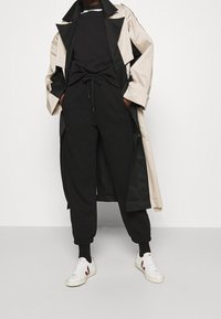 Trench coat oversized beige y negro, suéter negro, joggers negros con puños, zapatillas blancas con acentos rojos, diseño en capas y corte holgado.