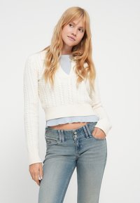 Κρεμ πουλόβερ με πλέξη καλώδιο, V-λαιμόκοψη και cropped σχεδιασμό, φορεμένο πάνω από ένα ανοιχτό γαλάζιο τοπ με βολάν, συνδυασμένο με ανοιχτό γαλάζιο τζιν.