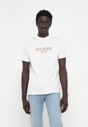 Camiseta blanca de algodón con un logo marrón texturizado que dice "GUESS EST. 1981" en el frente. Combinada con jeans de mezclilla azul claro.