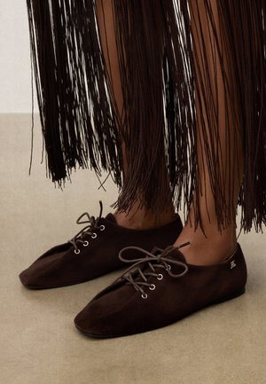 Chaussures en daim marron foncé à lacets portées avec une longue jupe à franges marron, debout sur un sol beige.