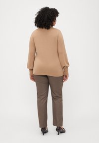 Pull en tricot beige avec une coupe décontractée et des manches longues, associé à un pantalon marron sur mesure et des talons en vernis foncé. Le matériau semble doux.