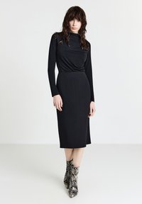 Donna con capelli effetto bagnato che indossa un vestito lungo nero a maniche lunghe e stivali a stampa serpente fino al ginocchio, su uno sfondo bianco uniforme.