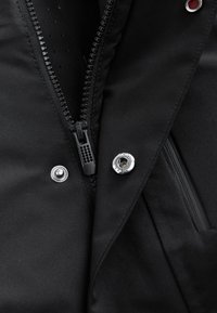 Sort isoleret jakke med frontlynlås, trykknapper og glat stoftstruktur med subtile perforeringer for åndbarhed.