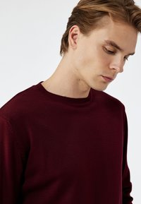 Burgundy neulepaita, jossa on pyöreä pääntie, pitkät hihat ja ribbaatut hihansuut. Tasainen rakenne, jossa on tasainen ommel ja minimaalinen muotoilu.