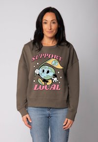 Bruine sweatshirt met een cartoon-wereldbol die een gele vlag vasthoudt, met roze tekst "SUPPORT LOCAL." Casual pasvorm, zachte textuur.