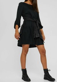 Vestido negro con mangas tres cuartos, cinturón en la cintura, falda acampanada y tela texturizada. Combinado con botas de tobillo negras con cordones.