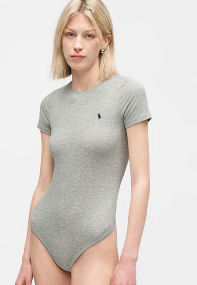 Νεαρή γυναίκα με ίσια ξανθά μαλλιά που φοράει εφαρμοστό γκρι κορμάκι με κοντά μανίκια και ένα μικρό ναυτικό λογότυπο στο στήθος.