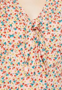 Lähikuva kukkakankaasta, jossa on pieniä punaisia, keltaisia ja turkooseja kukkia vaaleanpunaisella taustalla, kauluksessa solmittu solmu.