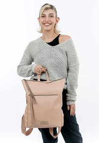 Zaino in pelle rosa chiaro con una tasca frontale con zip, doppi manici superiori e spallacci regolabili. Presenta linee pulite e hardware minimale.