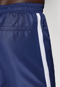 Mørkeblå treningsshorts med teksturert overflate, med hvite sidestreker og en baklomme. Elastisk midjebånd for justerbar passform.