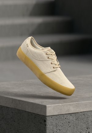 Šviesiai rudos spalvos drobiniai sportbačiai su šviesiai rudos gumos padais ir raišteliais, sklendžiančiais virš pilkos betono pakylos, su laiptais neryškioje fone.