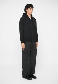 Mlad muškarac stoji u crnoj majici sa patentnim zatvaračem, ležernim crnim cargo hlačama sa velikim džepovima i crnim cipelama, na bijeloj pozadini.