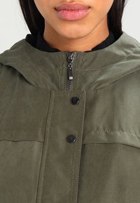 Chaqueta verde oliva con cierre frontal, botones a presión y capucha. El material parece suave con detalles de costura visibles.